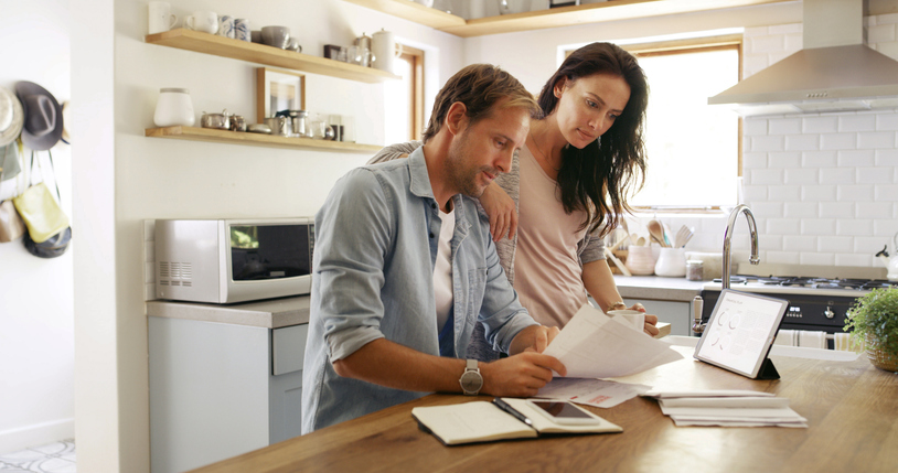 A couple reviews paperwork at a kitchen counter, surrounded by modern appliances and open shelves. They appear focused and engaged, conveying a cooperative tone.